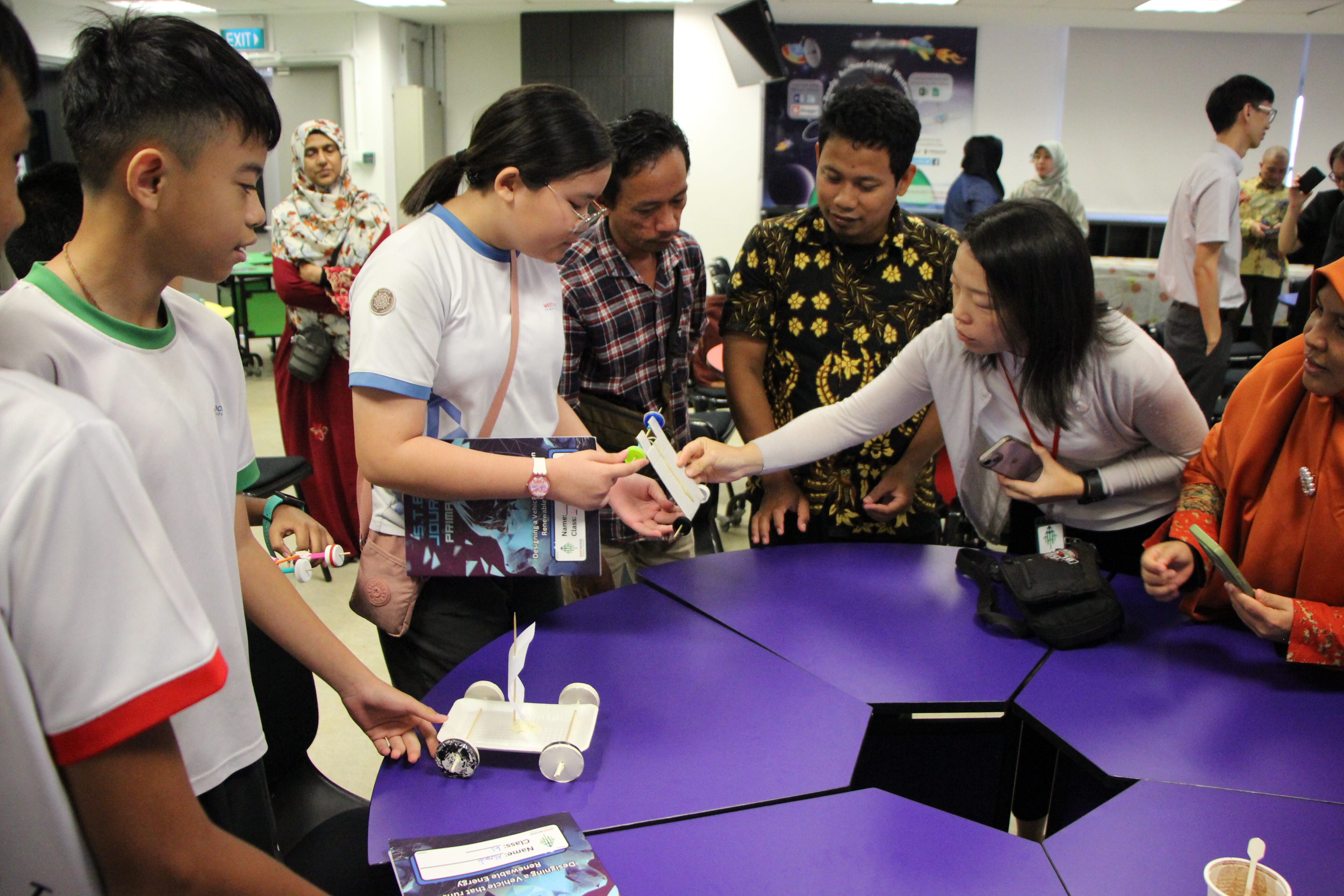 Students and adults gather around a purple hexagonal table examining a small model vehicle at a science event.
